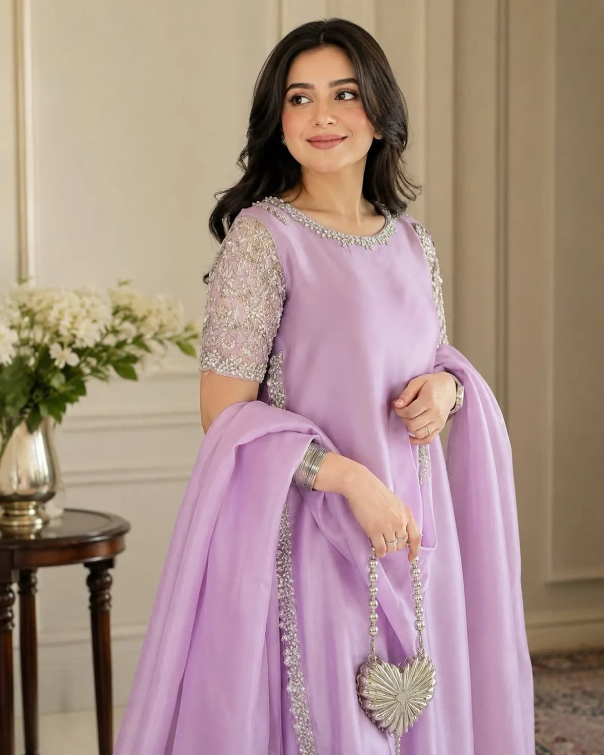 A woman models a lavender purple traditional Indian outfit with silver embellished embroidery on the bodice and sleeves, holding a matching purple dupatta and a silver heart-shaped handbag in an elegant indoor setting.
