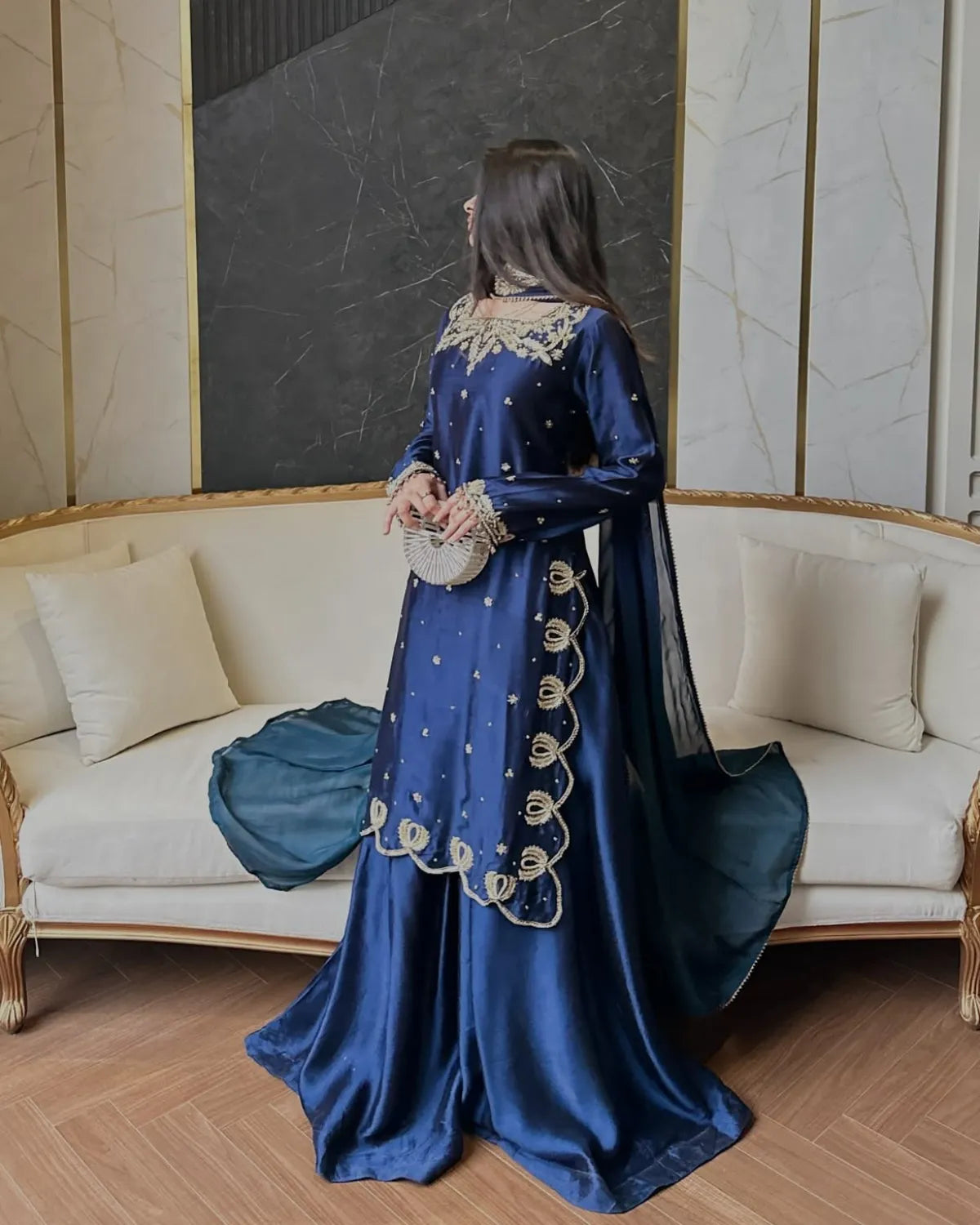 A woman stands in a furnished room wearing a long royal blue traditional outfit with gold embroidery and a matching sheer draped scarf. The setting includes a cream-colored sofa, wood flooring, and a dark wall panel.