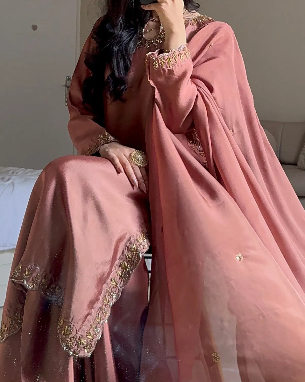 A woman seated indoors wearing a dusty pink traditional outfit with a matching sheer dupatta and ornate gold embroidery along the neckline, sleeve cuffs, and hem.