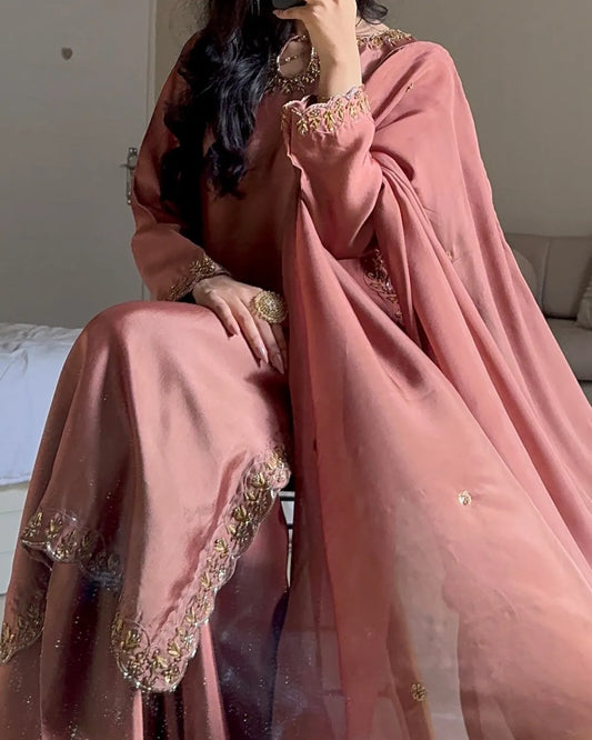 A woman seated indoors wearing a dusty pink traditional outfit with a matching sheer dupatta and ornate gold embroidery along the neckline, sleeve cuffs, and hem.
