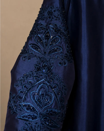 Close-up of a navy blue satin garment sleeve featuring intricate floral embroidery and beadwork, set against a solid beige background.
