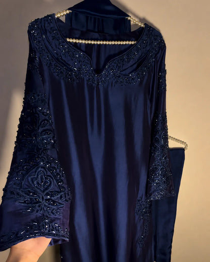 A navy blue satin caftan with intricate beadwork and embroidery on the neckline and sleeves, hanging on a hanger against a plain beige wall.