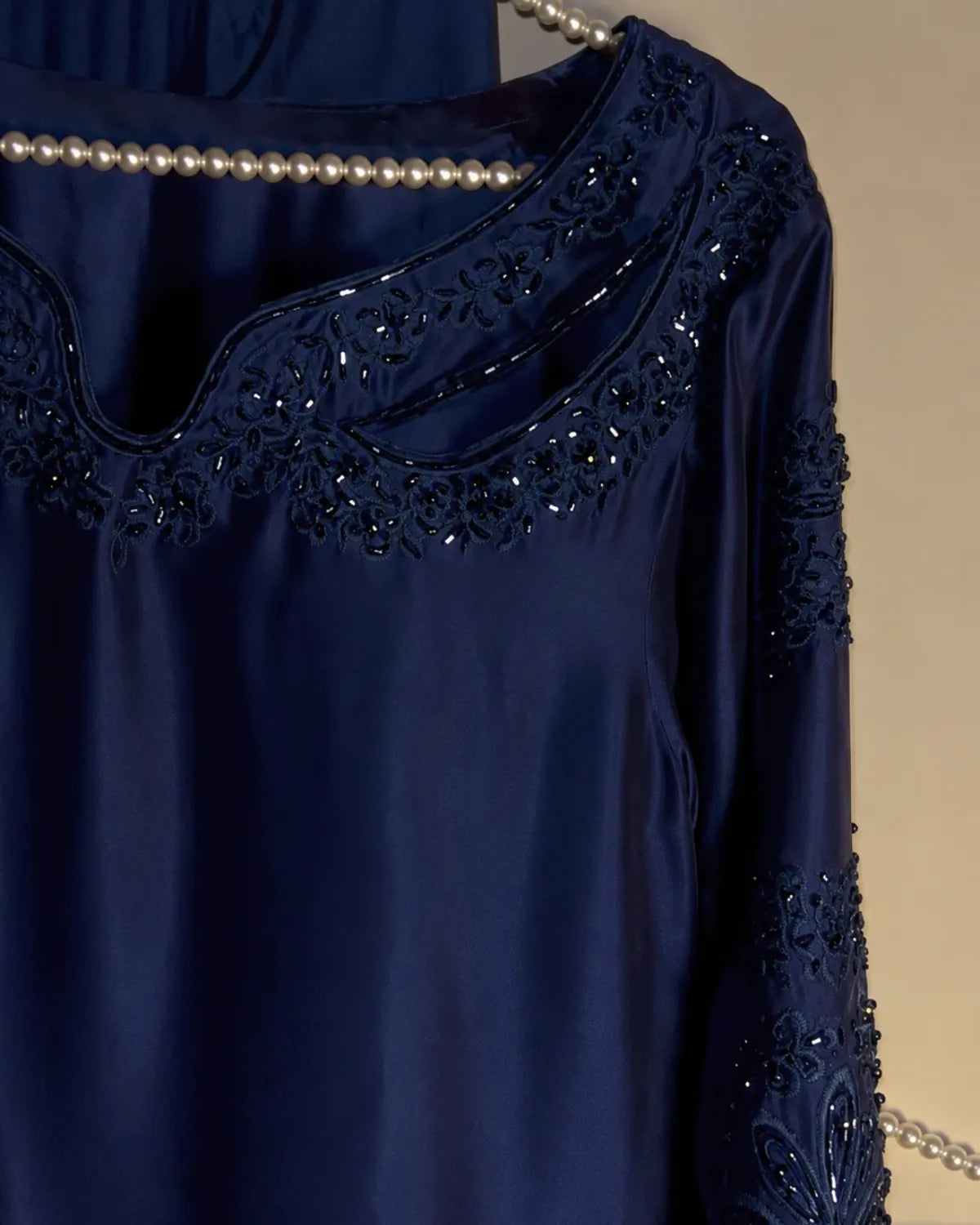 A close-up of a navy blue satin nightgown or tunic with intricate floral lace embroidery and sequined detailing around the scoop neckline and on the sleeve, styled with a pearl necklace against a neutral beige background.