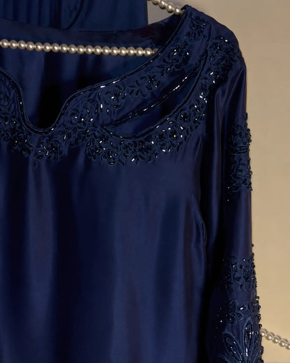 A close-up of a navy blue satin nightgown or tunic with intricate floral lace embroidery and sequined detailing around the scoop neckline and on the sleeve, styled with a pearl necklace against a neutral beige background.