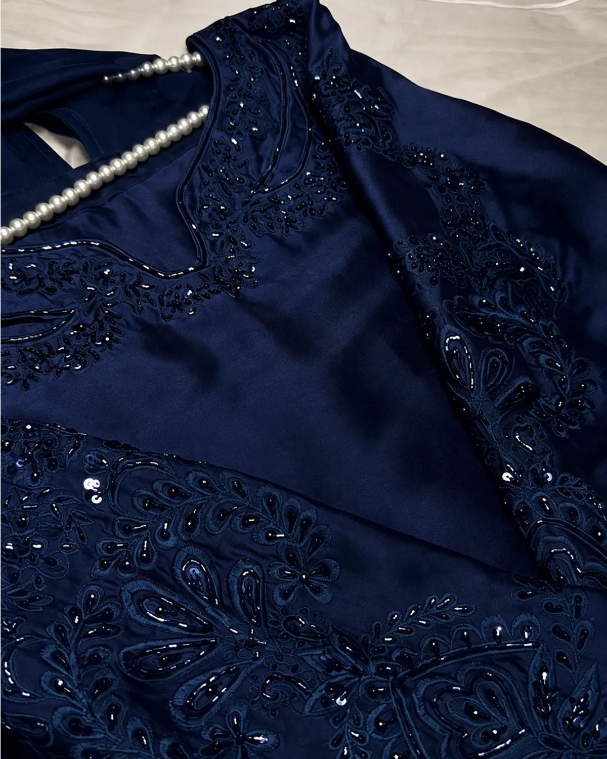 A close-up of a navy blue satin garment with intricate black beadwork and sequin embroidery, featuring a pearl necklace draped across the neckline.