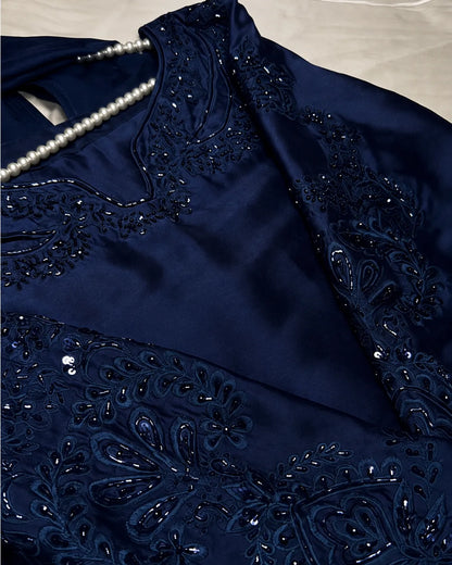 A close-up of a navy blue satin garment with intricate black beadwork and sequin embroidery, featuring a pearl necklace draped across the neckline.