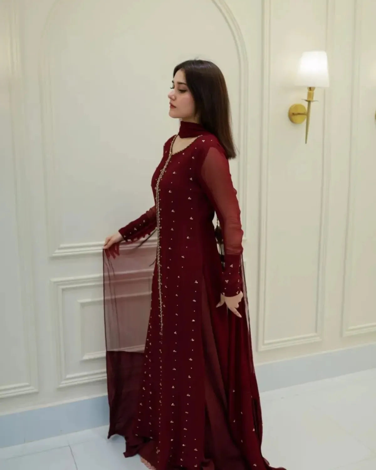 A woman wearing a deep maroon long-sleeve embroidered gown with sheer sleeves and a matching dupatta, standing in a cream-colored paneled room.