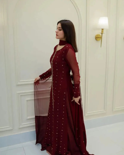 A woman wearing a deep maroon long-sleeve embroidered gown with sheer sleeves and a matching dupatta, standing in a cream-colored paneled room.