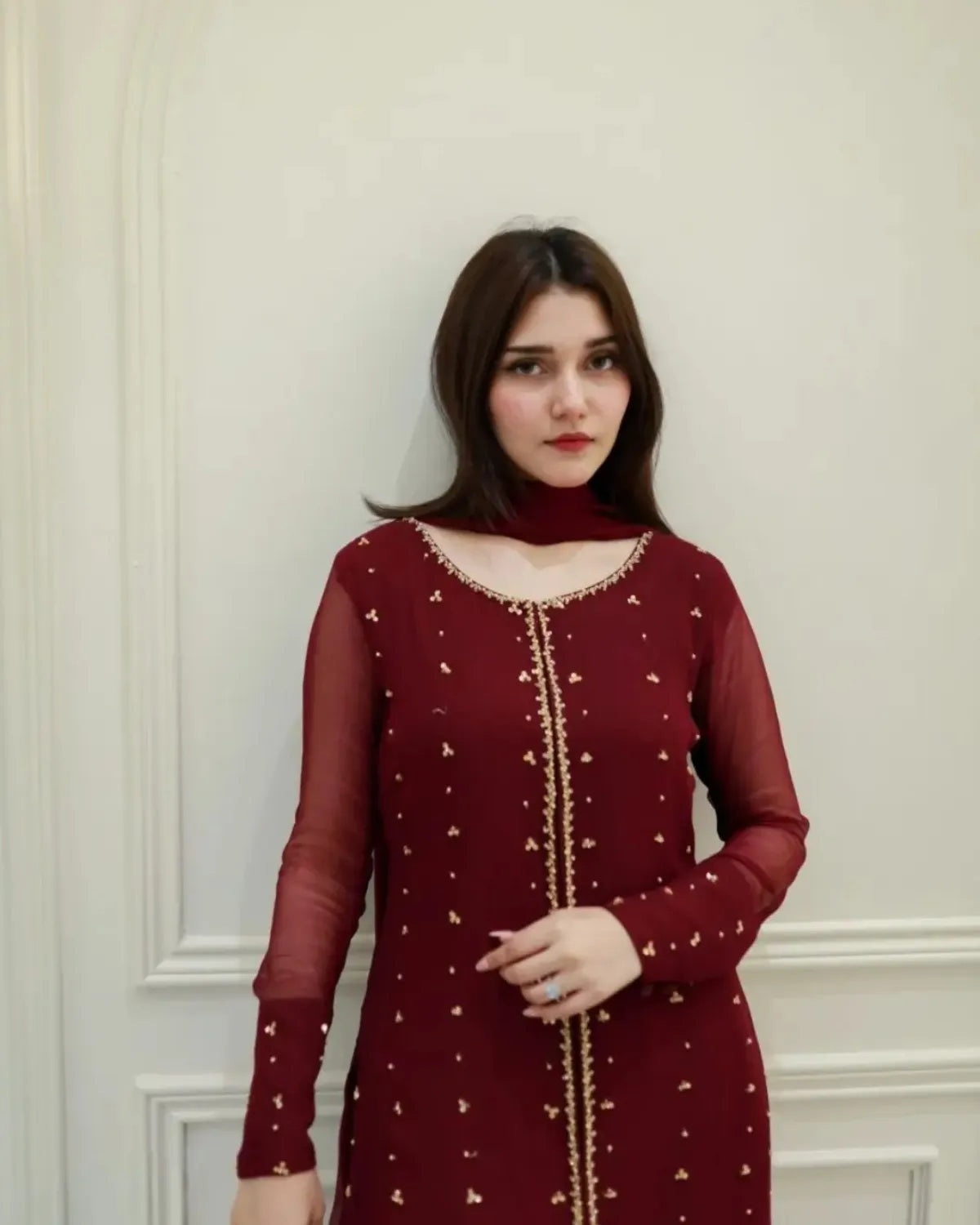 A woman wearing a deep maroon long-sleeve kurta with gold embroidery and a matching maroon neck scarf, standing against an off-white paneled wall.