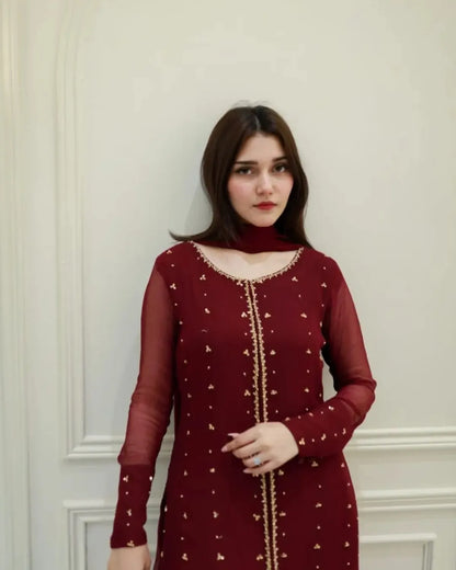 A woman wearing a deep maroon long-sleeve kurta with gold embroidery and a matching maroon neck scarf, standing against an off-white paneled wall.
