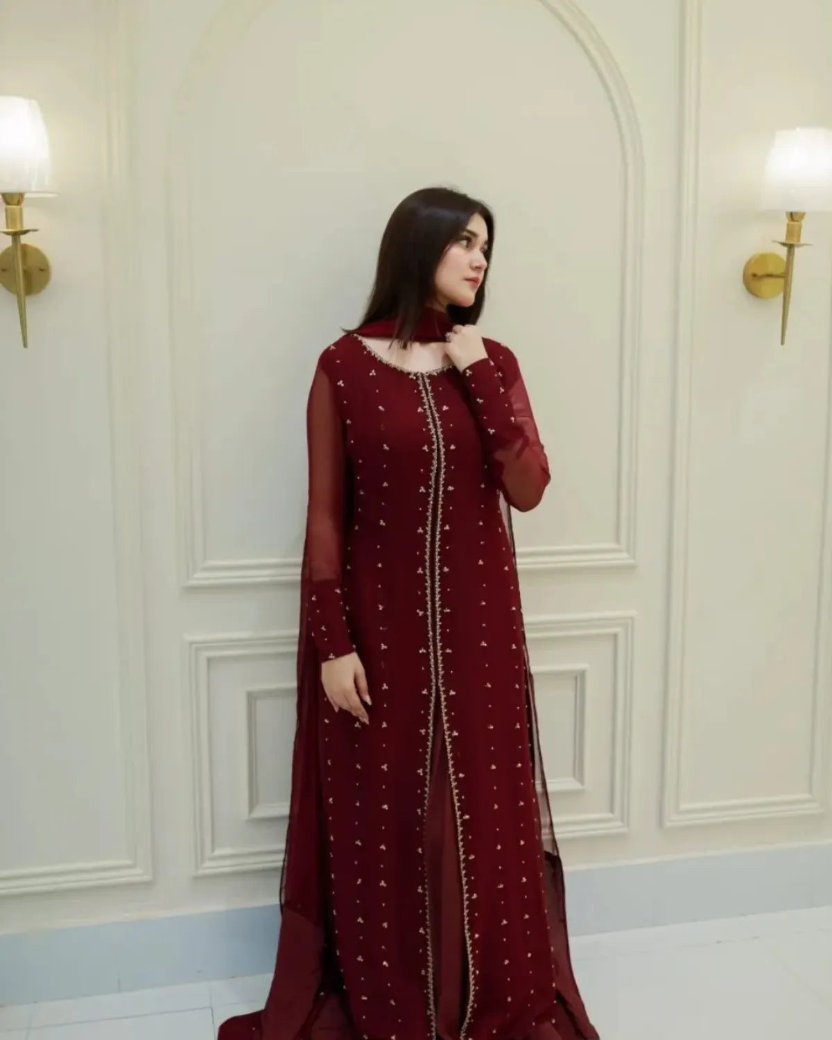 A woman wearing a deep maroon long gown with sheer sleeves and gold embellishments, standing in front of a cream-colored paneled wall with wall sconces.