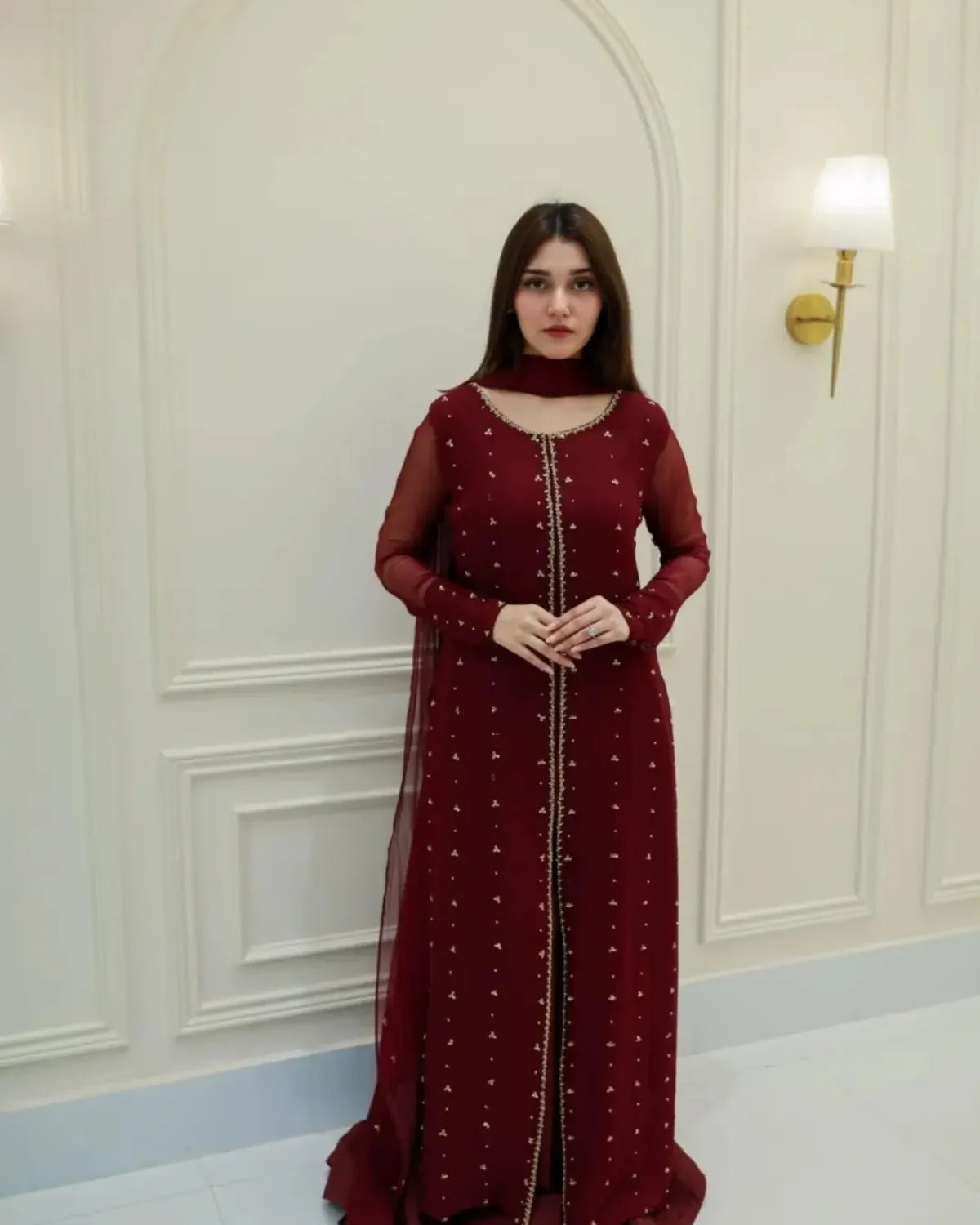 A woman wearing a deep maroon long-sleeve embroidered gown with a matching sheer dupatta, standing in front of a white paneled wall with a wall lamp.