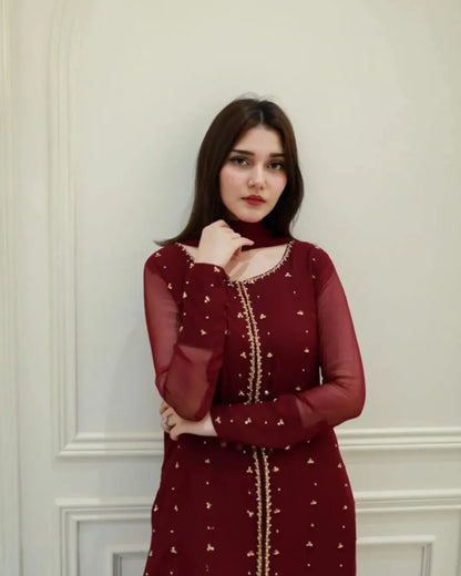 A woman wearing a deep maroon long-sleeve kurta with gold embroidery and a matching maroon neck scarf, standing against a white paneled wall.