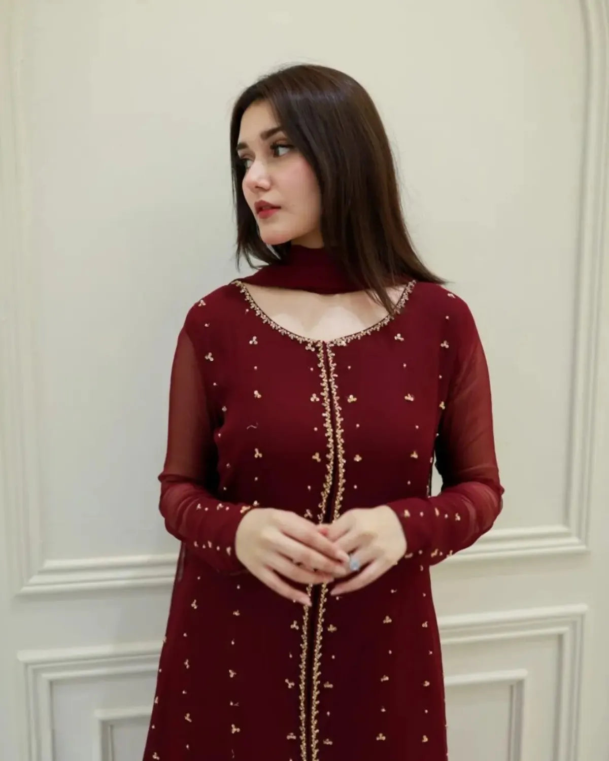 A woman wearing a deep maroon long-sleeve kurta with gold embroidery and a high neck, standing against a light cream paneled wall.