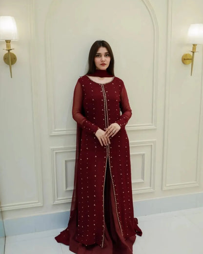 A woman stands indoors wearing a long maroon dress with sheer sleeves, a high neckline, and scattered gold embellishments. The background is a cream-colored paneled wall with two wall lamps.