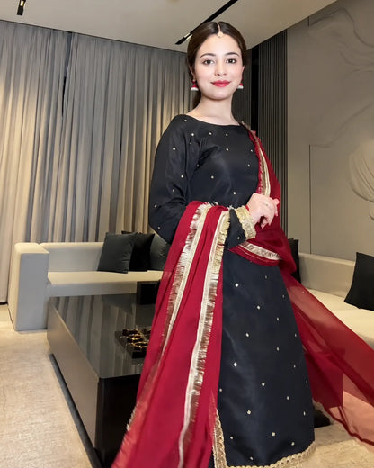 A woman wearing a black traditional outfit with gold dot embellishments and a red dupatta with gold border, standing in a modern living room setting.