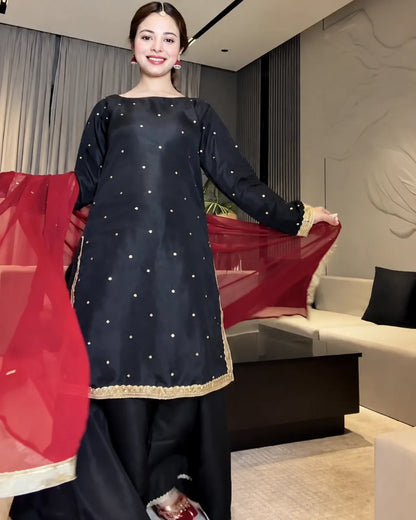 A woman modeling a black salwar kameez suit with gold zari embroidery and a red dupatta in a modern living room setting.