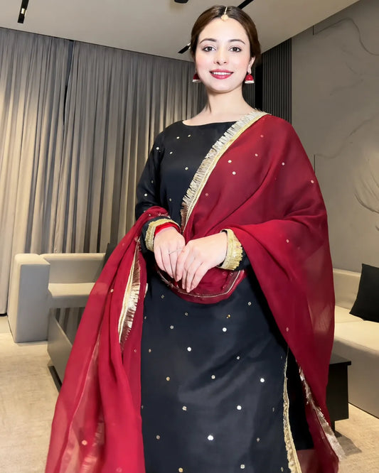 A woman wearing a black salwar kameez suit with gold dot embroidery and a deep red dupatta with gold border, standing in a modern living room setting.