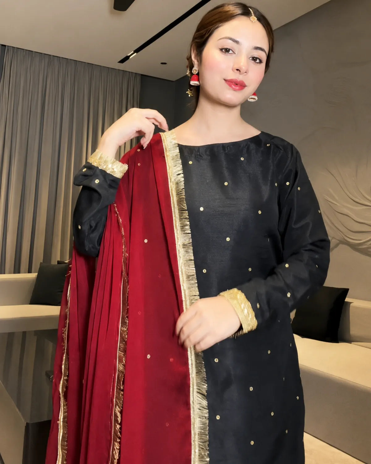 A woman modeling a black three-piece salwar kameez set with gold dot embroidery and a deep red dupatta featuring gold zari borders and gold accents.