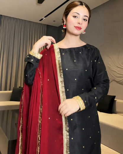 A woman modeling a black three-piece salwar kameez set with gold dot embroidery and a deep red dupatta featuring gold zari borders and gold accents.