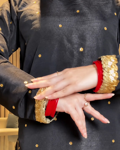 Close-up of a person wearing a black embroidered kurta with gold metallic dots and wide gold sequined cuffs, crossed over with red bangles on the hands.
