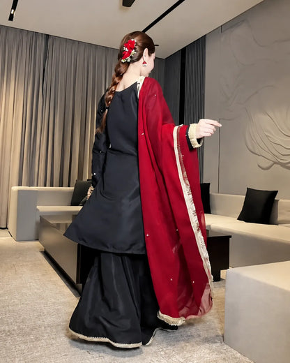 A woman wearing a black traditional outfit with a deep red dupatta draped over her shoulder, standing in a modern living room setting.