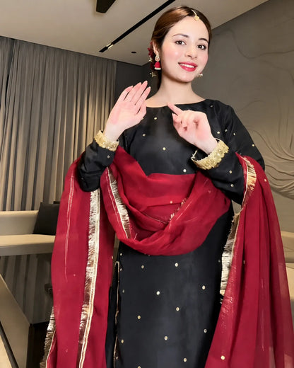 A woman wearing a black traditional outfit with gold dot embellishments and a deep red dupatta with gold zari borders, posing indoors with her hands raised in a greeting.