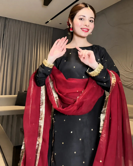 A woman wearing a black traditional outfit with gold dot embellishments and a deep red dupatta with gold zari borders, posing indoors with her hands raised in a greeting.