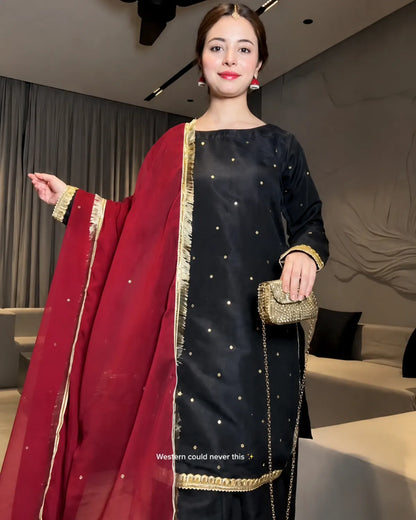 A woman models a black embroidered kurta set with a deep red dupatta and gold borders, holding a small gold clutch in an indoor setting.