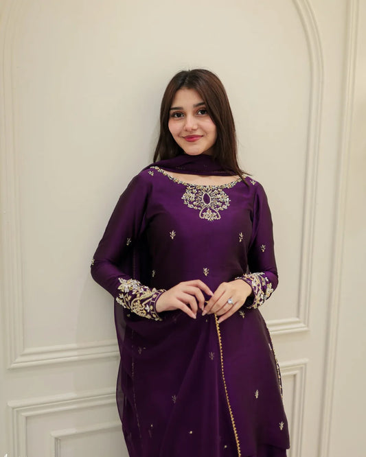 A woman wearing a deep purple traditional kurta with ornate silver and gold embroidery on the neckline and cuffs, standing against a cream-colored paneled wall.