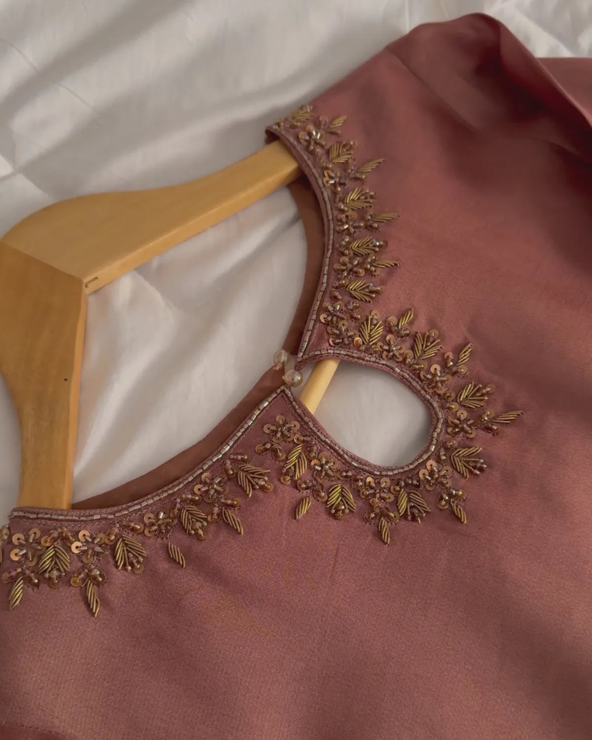 A close-up of a dusty rose pink blouse on a wooden hanger, featuring intricate metallic gold embroidery around the neckline.