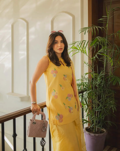 A woman stands on an indoor staircase wearing a bright yellow sleeveless embroidered dress or kurta set with floral motifs. She is holding a pink handbag and has sunglasses on her head, with a large potted plant and cream-colored walls in the background.