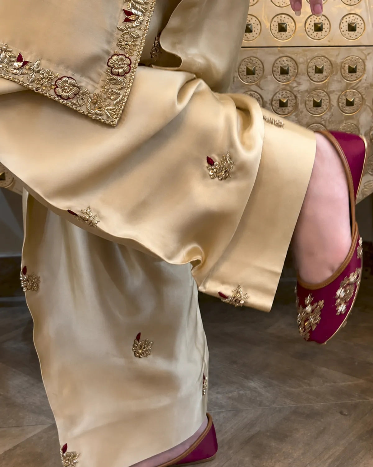 A close-up of a person wearing a cream satin traditional outfit with gold embroidery and magenta flat shoes with gold embellishments, standing in front of a decorative gold wall.