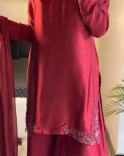 A person wearing a deep red traditional outfit with matching draped fabric, featuring silver embroidered detailing near the hem, shown indoors.