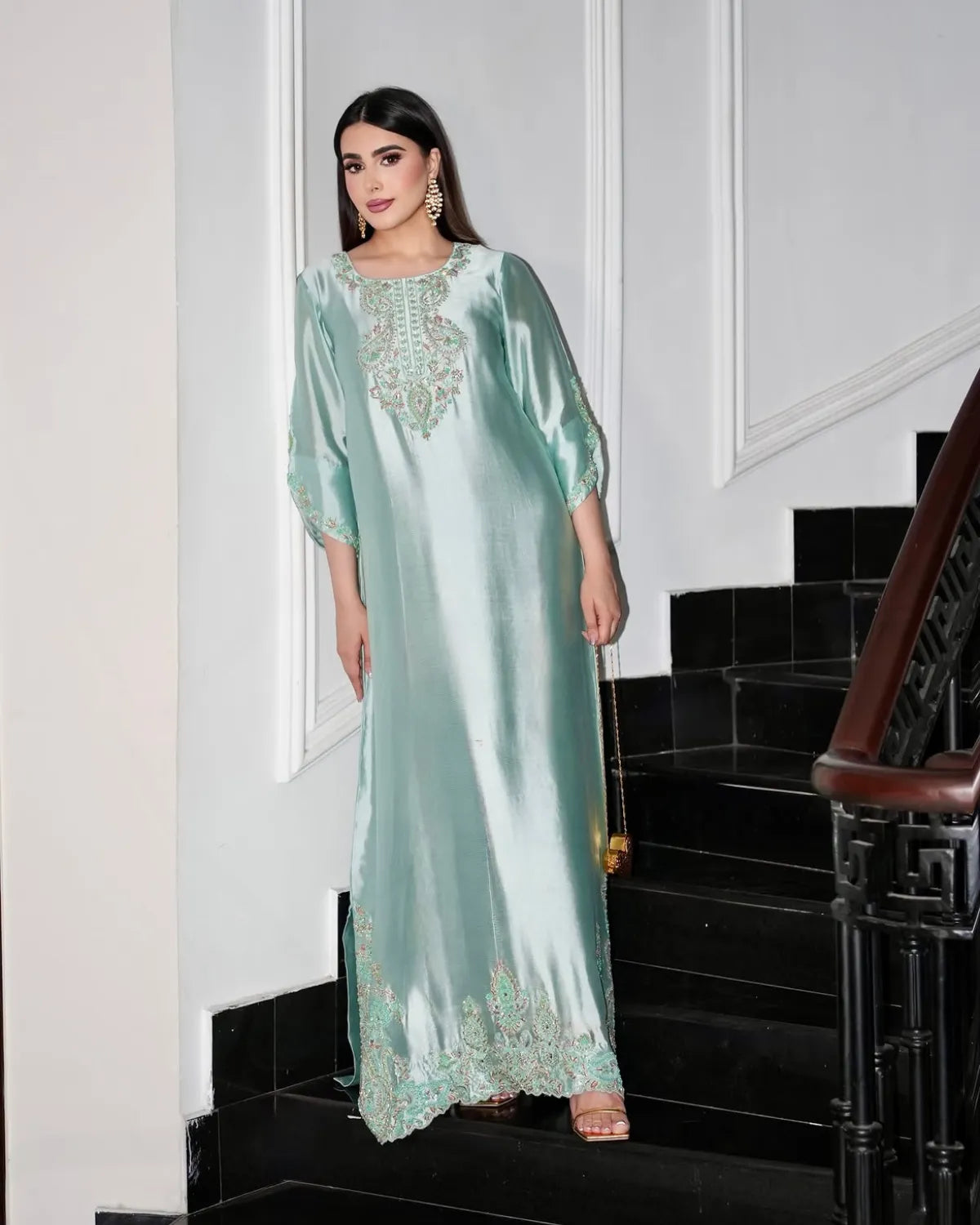 A woman models a mint green satin kaftan dress with intricate gold embroidery, standing on black stairs in a white-walled hallway.