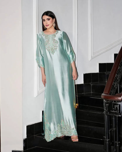 A woman models a mint green satin kaftan dress with intricate gold embroidery, standing on black stairs in a white-walled hallway.