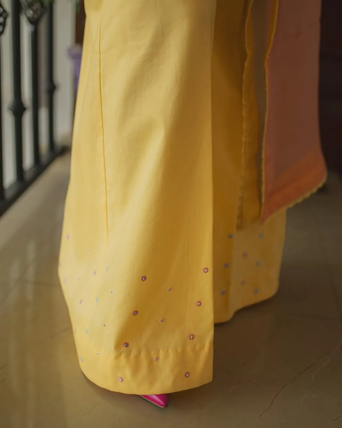 Close-up of a person's lower legs wearing wide-leg yellow palazzo pants with small colorful embroidered dots, paired with a peach-pink dupatta, standing on a tiled floor.