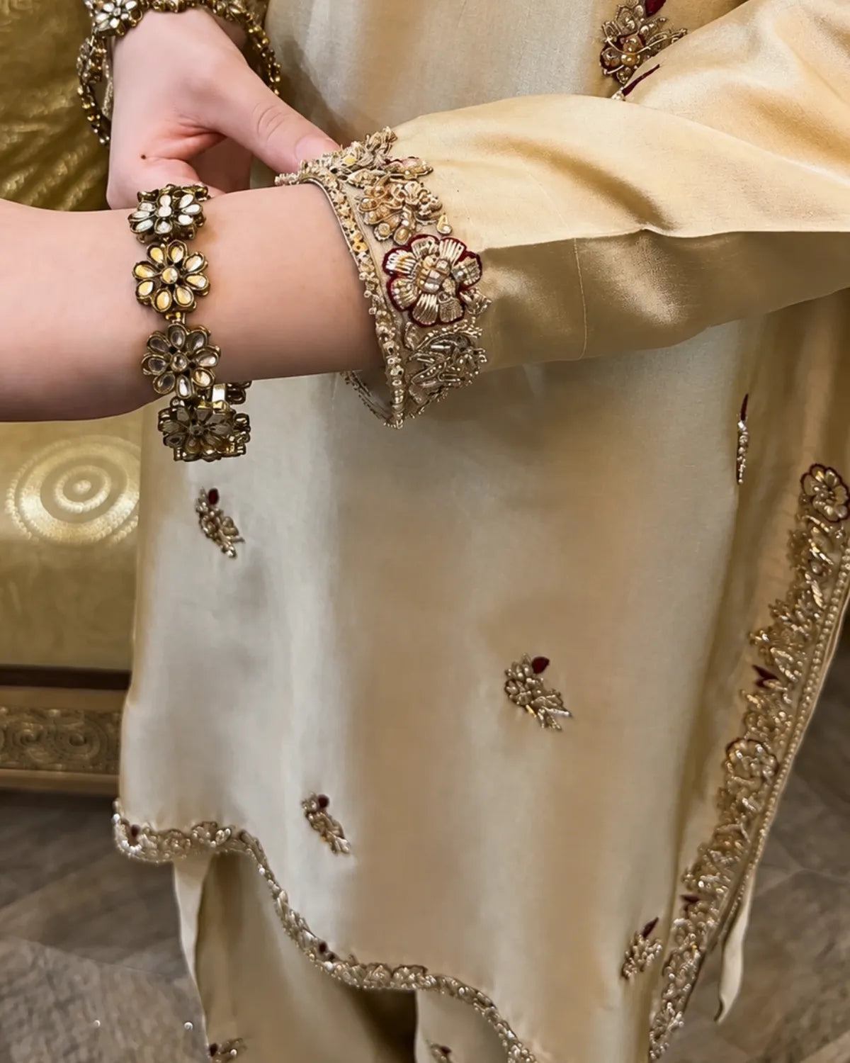 Close-up of a person wearing an ornate cream-colored traditional outfit with gold embroidery and embellished wrist bracelets.