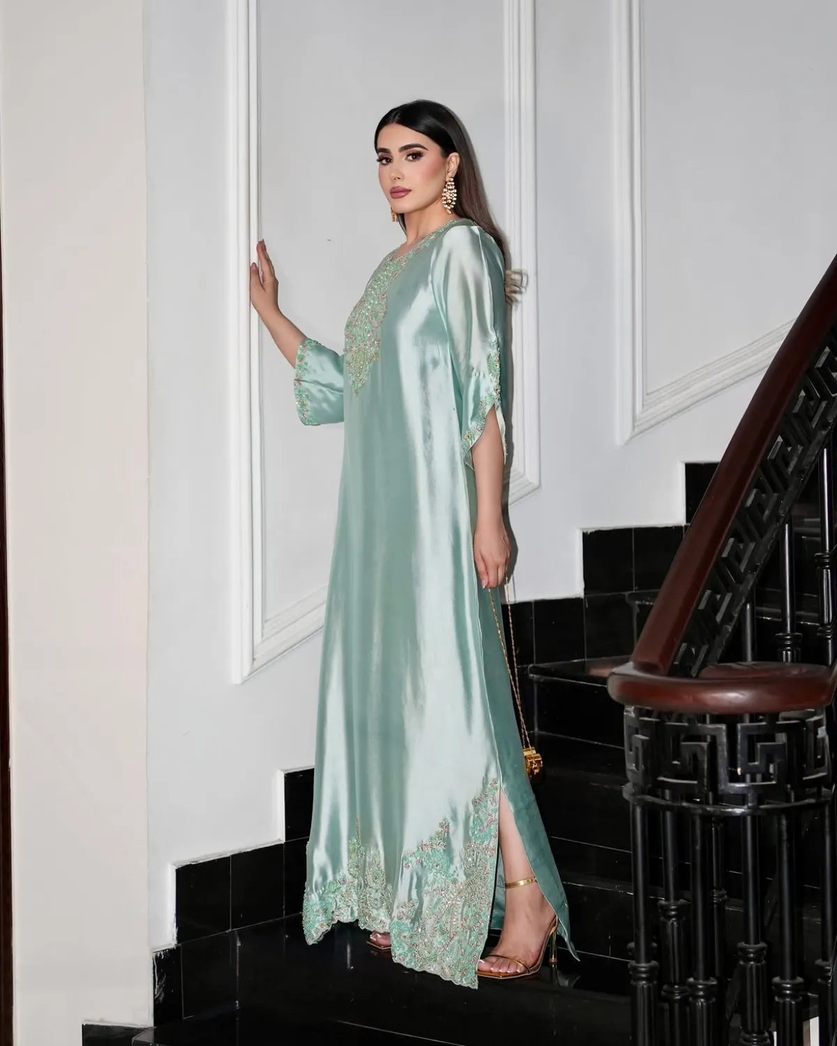 A woman models a floor-length mint green satin caftan with intricate gold embroidery on the neckline, sleeves, and hem, standing on a black staircase.