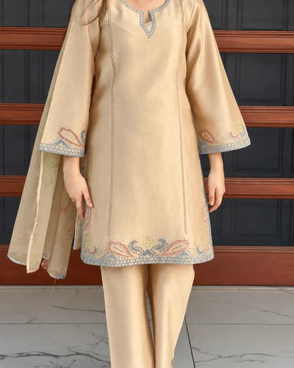 A model wearing a beige three-piece traditional South Asian outfit, featuring a straight kurta with blue and pink embroidered borders, matching trousers, and a coordinating dupatta, set against a dark paneled background.