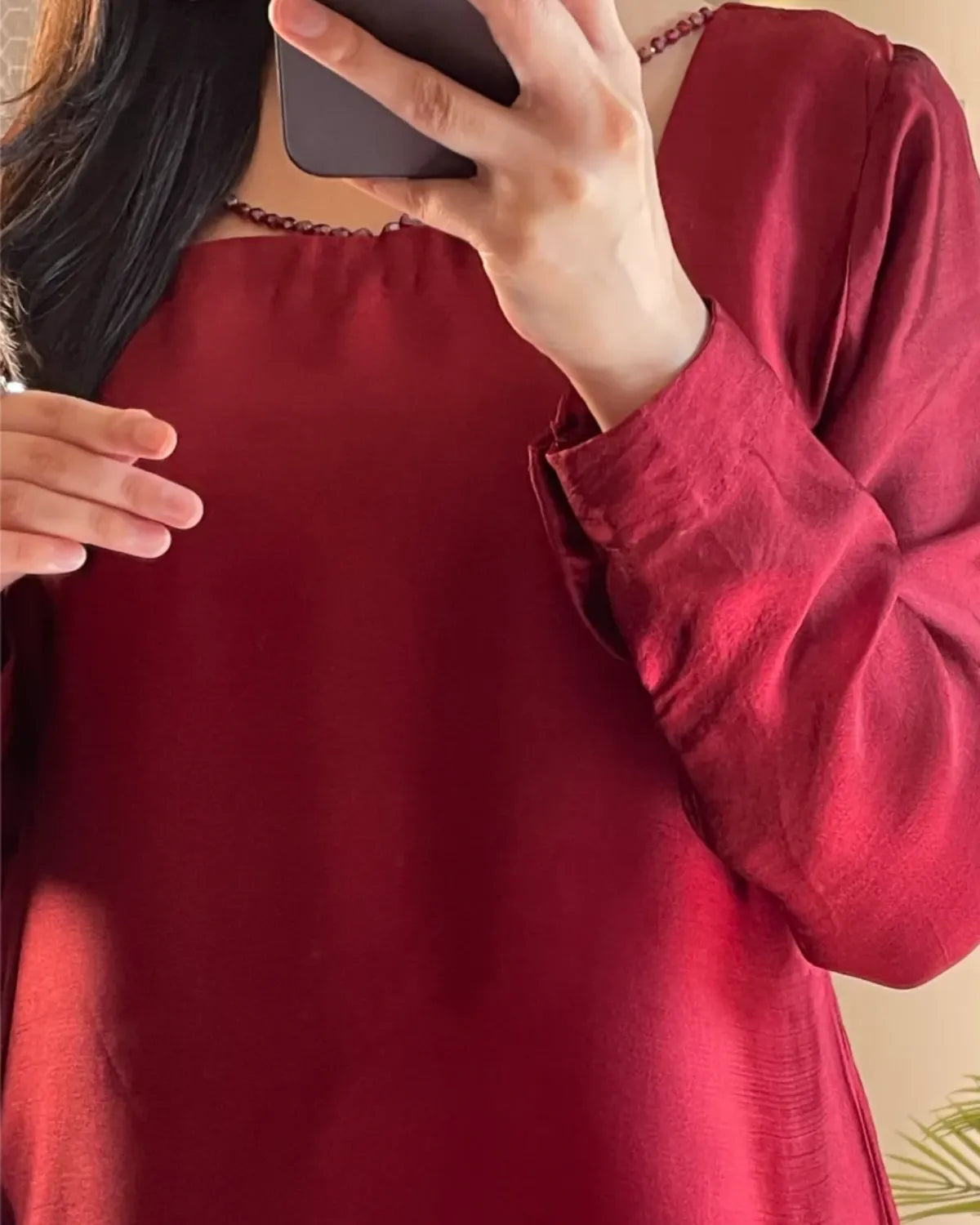 A close-up of a person wearing a long-sleeved deep red satin or silk top, taking a mirror selfie with a phone.
