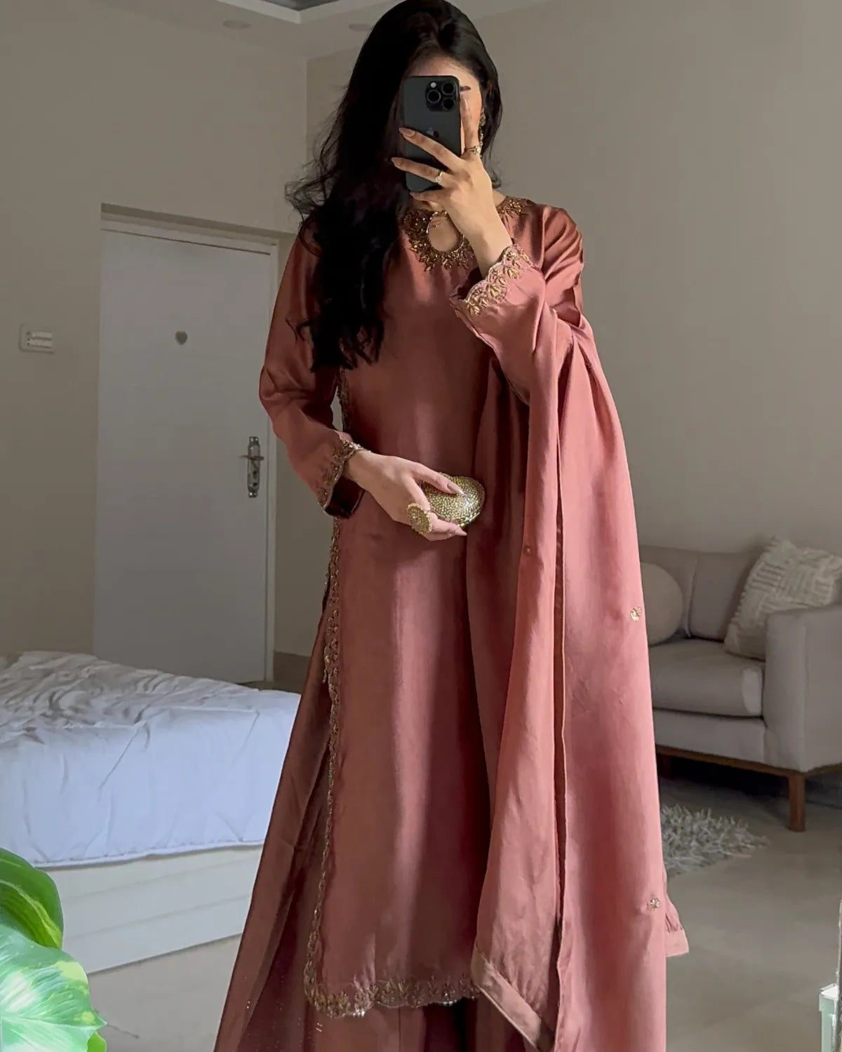 A woman takes a mirror selfie in a bedroom setting, wearing a dusty rose pink traditional outfit with gold embroidery and a matching draped scarf.