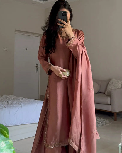A woman takes a mirror selfie in a bedroom setting, wearing a dusty rose pink traditional outfit with gold embroidery and a matching draped scarf.