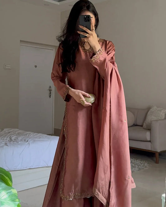 A woman takes a mirror selfie in a bedroom setting, wearing a dusty rose pink traditional outfit with gold embroidery and a matching draped scarf.