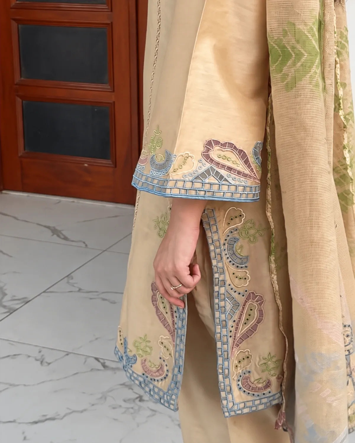 A close-up of a person wearing a beige traditional outfit with ornate multicolor paisley embroidery in blue, purple, and green, standing in front of a wooden door and marble floor.