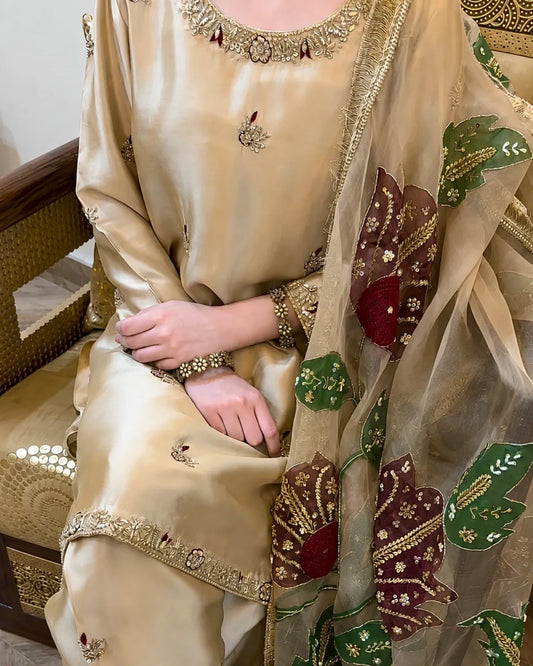 A close-up of a person wearing a beige satin traditional outfit with gold embroidery and a sheer dupatta featuring large green and burgundy floral motifs with gold detailing.