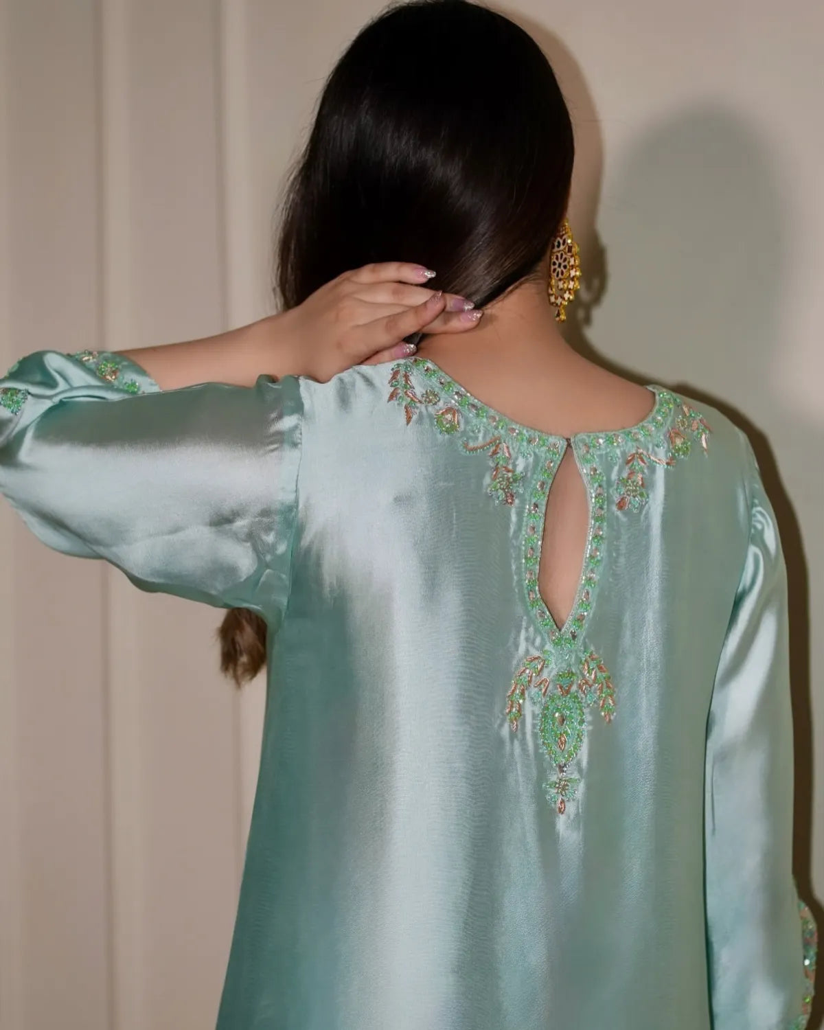 A woman shown from the back wearing a mint green satin tunic with intricate multicolor embroidery around the open back neckline. She has long dark hair and is wearing gold earrings against a neutral beige background.