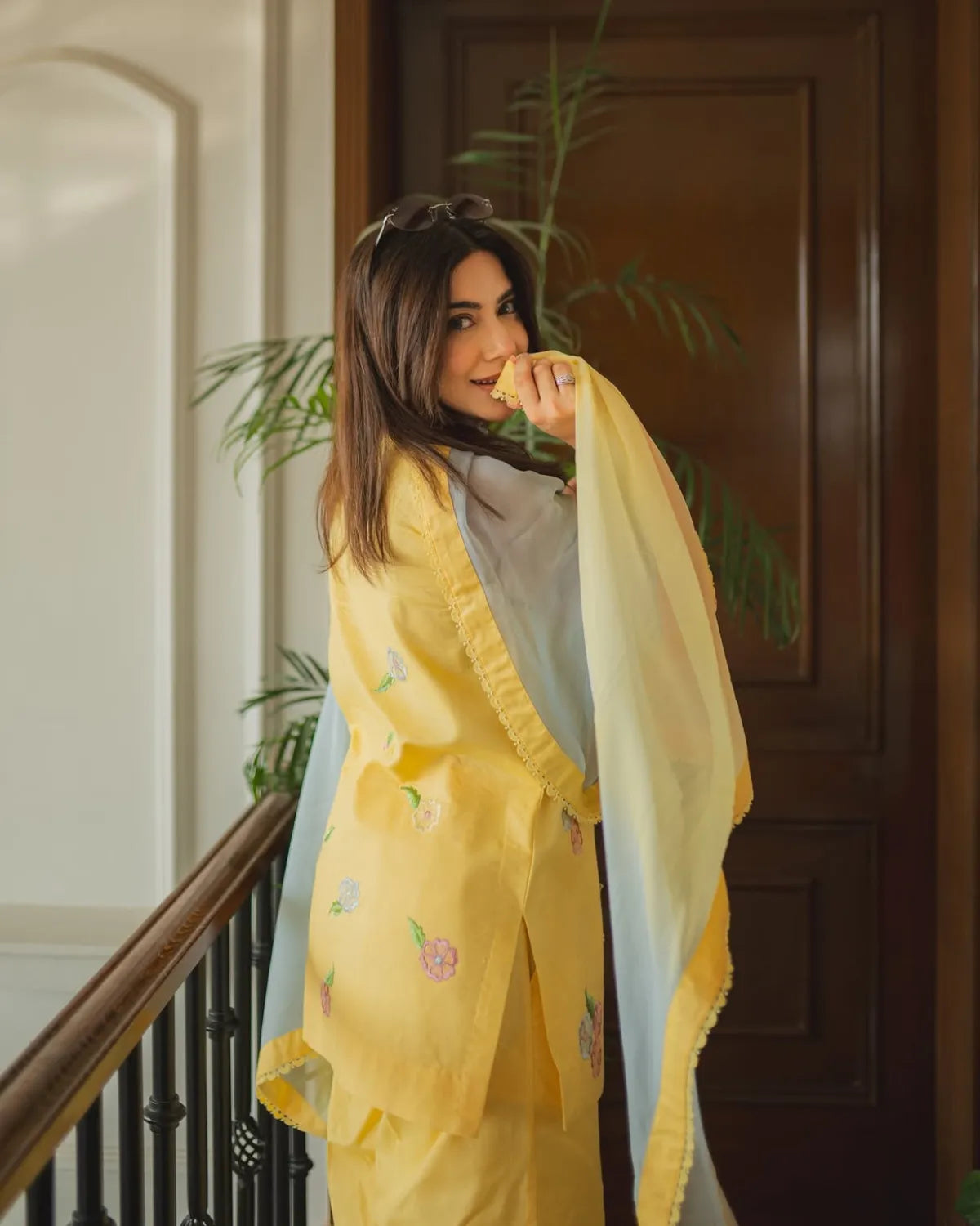 A woman wearing a yellow embroidered traditional outfit with a light blue dupatta, standing indoors near a wooden door and staircase railing.