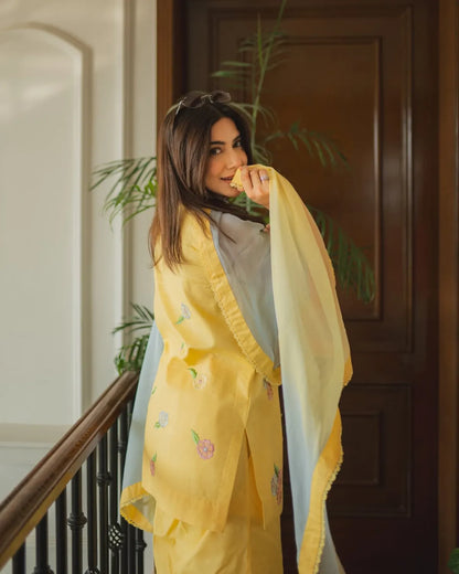 A woman wearing a yellow embroidered traditional outfit with a light blue dupatta, standing indoors near a wooden door and staircase railing.