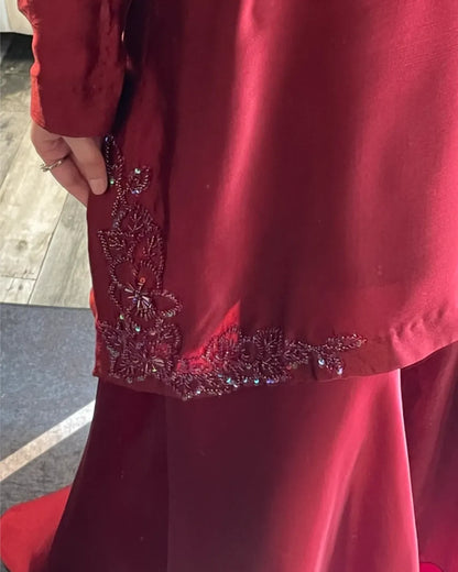 A close-up of a deep red satin-like garment with ornate embroidered detailing and sequined embellishments near the hem, worn by a person whose hand is visible holding the fabric.